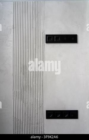 Mur intérieur minimaliste moderne avec prises et interrupteurs électriques noirs, panneau de plâtre texturé vertical décoratif et style béton gris clair Banque D'Images