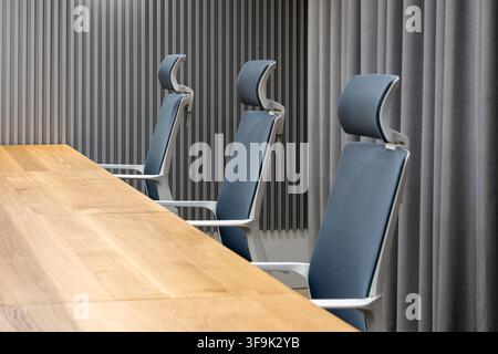 Salle de réunion de bureau moderne avec des chaises bleues ergonomiques, une grande table de conférence en bois naturel, des panneaux acoustiques muraux et des rideaux gris dans un intérieur de luxe Banque D'Images
