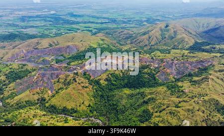 Vue aérienne d'une ferme solaire à grande échelle intégrée dans des collines verdoyantes ondulantes, avec des rangées de panneaux photovoltaïques et des routes d'accès à Aguilar, Ilocos Banque D'Images