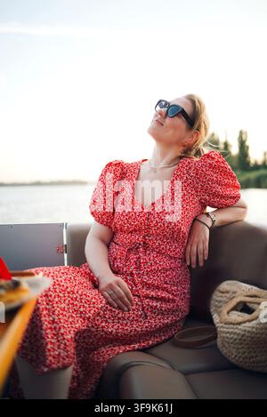 Femme en robe fleurie rouge profitant d'un moment de détente sur un bateau, avec de l'eau et des arbres en arrière-plan sous un ciel dégagé Banque D'Images