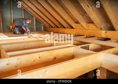 Une personne est dans le grenier en train de travailler sur une charpente en bois. L'espace a des poutres apparentes et l'ouvrier utilise des outils pour fixer la structure. La lumière traverse Banque D'Images