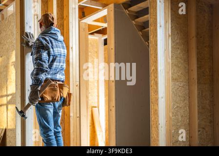 Un ouvrier se tient à côté de châssis en bois dans une maison en construction. L'ouvrier tient un marteau et porte des gants. L'espace montre des poutres en bois et des travaux inachevés Banque D'Images