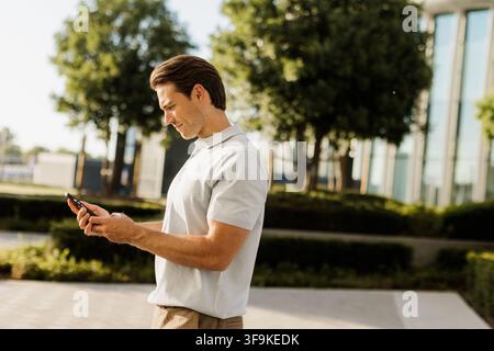 Un jeune homme se tient dans un parc verdoyant, absorbé par son smartphone alors que le soleil brille haut dans le ciel. Banque D'Images
