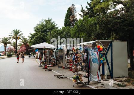 Bar, Monténégro – 26 juin 2026. Des kiosques à souvenirs exposent des jouets, des articles de plage et des cadeaux le long d'une promenade maritime bordée d'arbres avec des piétons. Banque D'Images