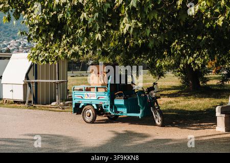 Bar, Monténégro - 26 juin 2026. Un véhicule utilitaire municipal à trois roues est garé sous un arbre feuillu près de la promenade côtière. Banque D'Images