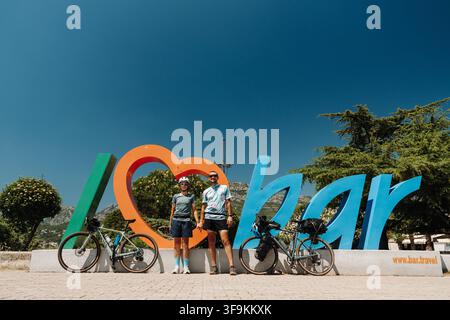Bar, Monténégro – 26 juin 2026. Un homme et une femme cyclistes posent avec des vélos de tourisme chargés devant l’enseigne colorée « I Love Bar ». Banque D'Images
