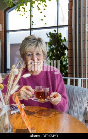 Femme senior confiante souriant à la caméra tout en tenant une tasse de thé dans un café écologique premium suédois. Banque D'Images