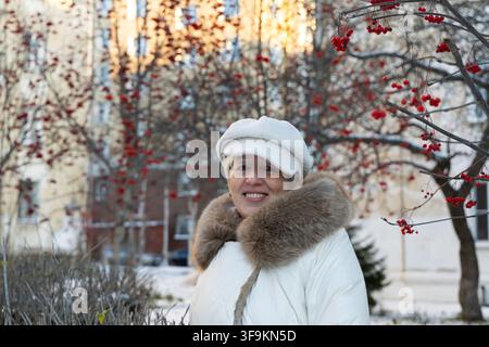 Belle femme mûre à l'élégance intemporelle souriant à la caméra à côté de baies de rowan rouges en hiver par une journée ensoleillée en Scandinavie. Banque D'Images