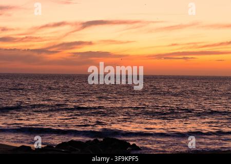 Le coucher de soleil peint l'océan dans des teintes chaudes d'orange et de rose. Des vagues douces clapotent contre la côte rocheuse. Le ciel reflète des tons ardents sur les eaux calmes. Scène évoque Banque D'Images
