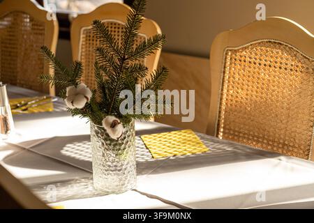 Centre de table avec des branches de sapin et des fleurs de coton dans un vase en verre texturé sur une table à manger avec des chaises en osier, illuminé par la lumière du soleil dans un restaurant Banque D'Images