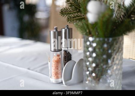 Moulins à sel et poivre en acier inoxydable et en verre avec du sel rose de l'Himalaya et des grains de poivre à côté d'un porte-serviettes sur une table couverte d'une nappe blanche. Banque D'Images