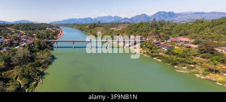 Une vue aérienne montre un pont traversant la rivière Nam Kading entre les hameaux riverains et les collines boisées, avec une chaîne de montagnes au loin. Banque D'Images