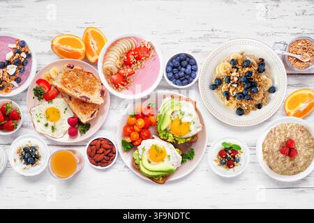 Scène de table pour un petit-déjeuner ou un brunch sain sur un fond en bois blanc. Vue de dessus. Toast à l'avocat, bols de smoothies, avoine, yaourt et une variété de nutriments Banque D'Images