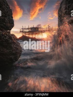 Vue du Trabocco Spezzacatena au coucher du soleil avec des vagues s'écrasant contre les rochers au premier plan et un ciel orange vif à Rocca San Giovanni, Province Banque D'Images
