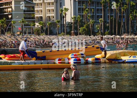 Les employés du resort préparent des bateaux bananes gonflables sur le ponton tandis qu'un couple de personnes âgées nage dans la mer devant un fond de plage bondée à Icmeler, en Turquie. Banque D'Images