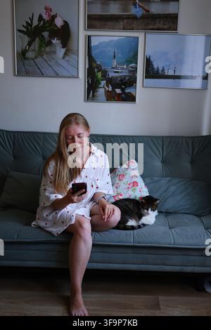 Jeune femme détendue utilisant un téléphone portable avec un chat domestique à proximité, technologie moderne et vie quotidienne à la maison. Banque D'Images