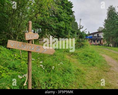 Un panneau routier rustique en bois se dresse dans un cadre rural verdoyant, avec deux panneaux peints à la main indiquant différentes villes. Le plus grand Banque D'Images