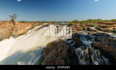 Vue en pose longue de larges cascades dévalant sur des rochers exposés aux chutes de Li Phi Somphamit, avec plusieurs chenaux sous un ciel bleu clair. Banque D'Images
