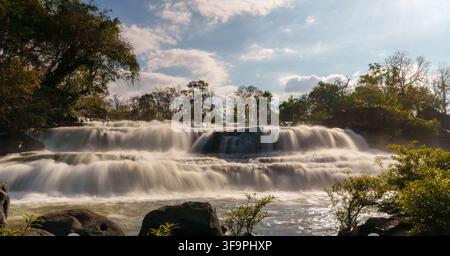 Une vue frontale large de la cascade de Tad Hang montre plusieurs niveaux rocheux, une brume rétroéclairée et des rochers au premier plan. Banque D'Images