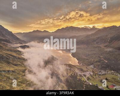 Vue aérienne de la vallée de l'Engadine avec un lac serein reflétant la lumière de l'heure dorée, entourée de montagnes couvertes de brume et de forêts luxuriantes dans les Grisons, S Banque D'Images
