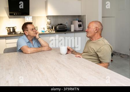 Un couple de personnes âgées est assis à une table de cuisine. La femme semble pensive en écoutant l'homme. Il tient une tasse à la main. Ils semblent avoir une conversation à la maison. Banque D'Images