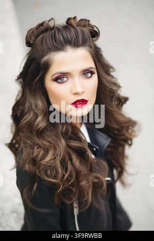 Portrait de belle femme avec de longs cheveux bouclés, rouge lèvres et maquillage parfait, qui marche à rue. Girl à la mode veste en cuir en lo Banque D'Images