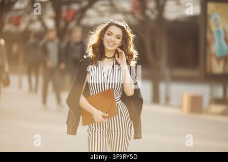 Belle femme brune dans un look printemps élégant, portant un costume rayé et une veste noire, tenant un sac à main rouge, fille souriante avec maquillage après la beauté Banque D'Images