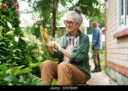 Sénior mettant des gants de jardinage dans le jardin de fleurs de l'arrière-cour Banque D'Images