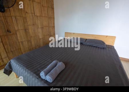 Une chambre avec un lit en bois, une parure de lit à rayures sombres, des oreillers et des serviettes roulées créant un intérieur rangé et reposant avec des meubles naturels et un style rustique chaleureux Banque D'Images