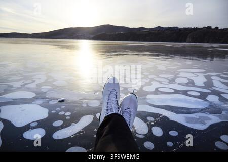 Jambes de femme en patins blancs sur une patinoire extérieure au coucher du soleil. Lac gelé Baïkal Banque D'Images