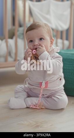 Petite fille en barboteuse rose jouant avec un hochet en bois et tendant la main vers des perles tout en étant assise sur un sol en bois près d'un berceau dans un bâtiment ; curiosité tranquille Banque D'Images