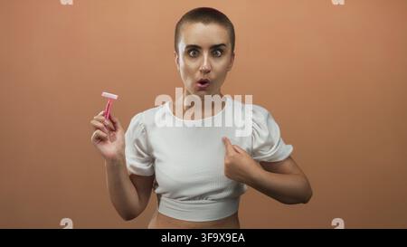 Femme aux cheveux courts tenant un rasoir jetable rose, pointe le doigt sur la poitrine tout en portant un haut blanc recadré en studio ; autonomisation. Banque D'Images