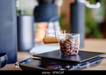 Grains de café torréfiés dans une tasse en verre transparent sur une balance numérique avant le broyage. Gros plan de café mesuré préparé pour une infusion manuelle à la maison. Banque D'Images