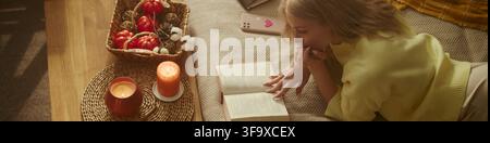Jeune femme caucasienne allongée sur le sol en train de lire un livre près de bougies allumées et de décorations automnales. Vue en hauteur d'une activité de loisirs confortable idéale pour les campagnes marketing saisonnières Banque D'Images