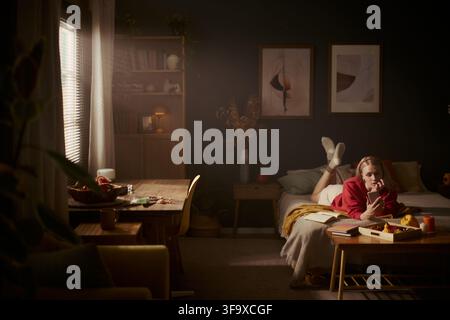 Jeune femme caucasienne allongée sur le lit et utilisant un smartphone dans une chambre confortable. L'image représente un mode de vie numérique et des loisirs à distance, adaptée au marketing d'applications mobiles Banque D'Images