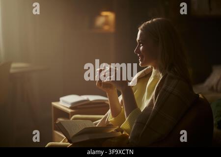 Jeune femme caucasienne assise dans un fauteuil, tenant une boisson chaude et souriant, profitant de son temps libre à la maison. L'image représente le bien-être mental et les soins personnels, idéale pour le marketing de style de vie Banque D'Images