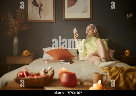 Jeune femme caucasienne portant des écouteurs, allongée dans un lit et utilisant un smartphone. L'image représente un mode de vie numérique et convient aux campagnes de bien-être mental et de loisirs à distance Banque D'Images