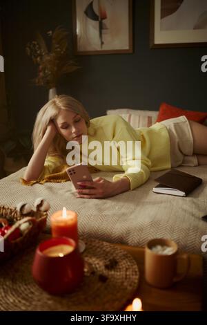 Image verticale d'une jeune femme caucasienne allongée sur un lit et utilisant un smartphone. L'image représente la vie numérique et la détente, adaptée au bien-être mental et au marketing de style de vie Banque D'Images
