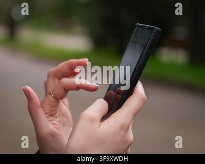 Les mains d'une personne tenant un smartphone noir, touchant l'écran réfléchissant avec un doigt, effectuant une action sur l'appareil mobile en extérieur Banque D'Images