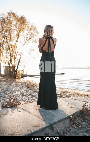 Femme élégante en robe noire longue debout au bord d'un lac au coucher du soleil. Photo d'archive haute résolution. Banque D'Images