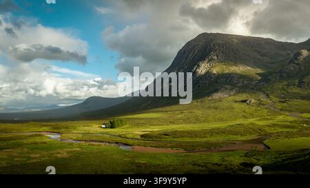 Vue aérienne de la vallée dramatique de Glencoe présentant des montagnes escarpées, une rivière sinueuse et une petite maison nichée dans le paysage verdoyant sous un nuage Banque D'Images
