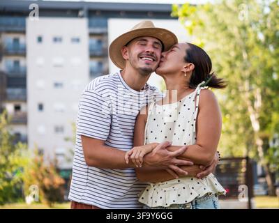 Homme et femme joyeux partageant une étreinte chaleureuse et un baiser entourés de verdure dans un parc urbain. Banque D'Images