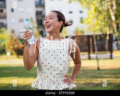 Femme adulte moyenne boit de l'eau dans le parc par une journée d'été ensoleillée. Elle regarde la caméra et sourit. Banque D'Images