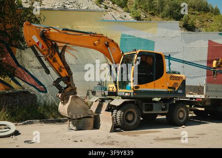 Excavatrice à roues imposante garée sur un chantier industriel, sa pelle positionnée au-dessus d'un conteneur métallique dans un environnement montagneux accidenté Banque D'Images