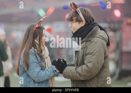 Jeune couple se tenant la main et souriant sur un marché de Noël portant des bandeaux de renne Banque D'Images