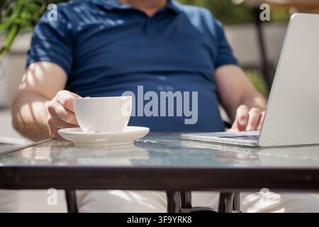 Homme caucasien travaillant à distance sur un ordinateur portable tout en atteignant son café dans un café en plein air ensoleillé, travail indépendant et mode de vie professionnel flexible. Banque D'Images