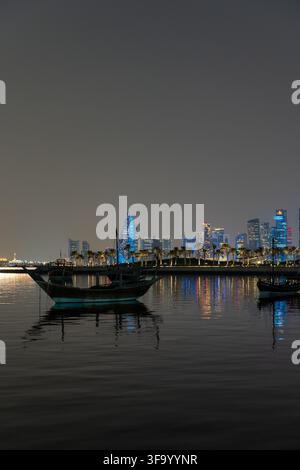 Doha, Qatar - 11 novembre 2025 : Une vue en composition verticale de bateaux dhow traditionnels amarrés sur une mer calme la nuit, mettant en vedette le moderne illuminé Banque D'Images