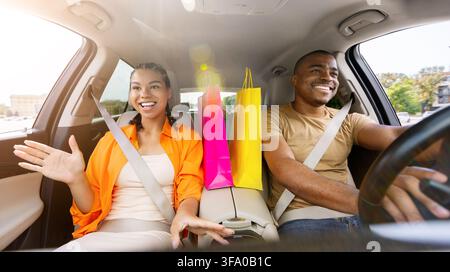 Couple noir joyeux voyageant en voiture avec des sacs de courses colorés entre eux Banque D'Images