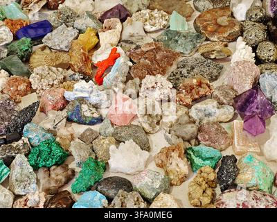 Collection colorée de pierres précieuses brutes et minéraux variés. Des cristaux aux couleurs vives sont dispersés sur une surface sablonneuse. Les pierres varient en forme, teinte et Banque D'Images