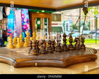 Jeu d'échecs avec pièces en bois sur plateau sculpté. L'arrière-plan montre une vitrine colorée. Scène de rue en plein air avec voitures et bâtiments. Éclairage chaud met en valeur Banque D'Images